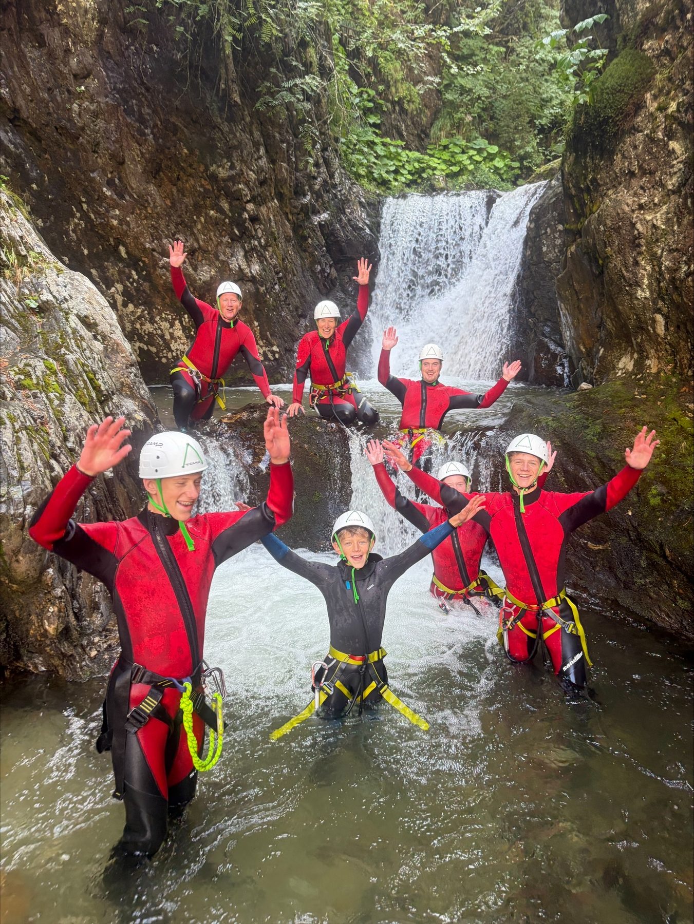Gruppe in Neoprenanzügen steht mit erhobenen Armen vor einem Wasserfall