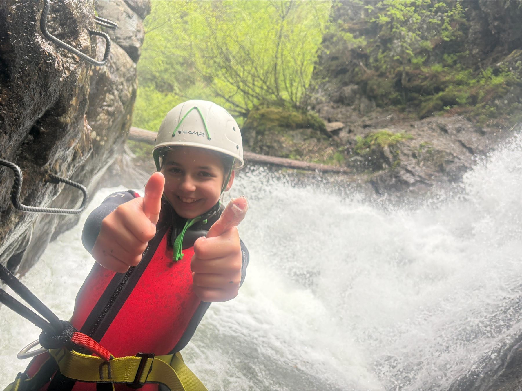 Kind mit Helm und Schwimmweste zeigt vor einem Wasserfall beide Daumen nach oben