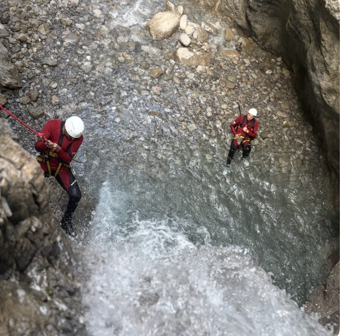 Person seilt sich an einer nassen Felswand ab, eine zweite wartet im Flussbett